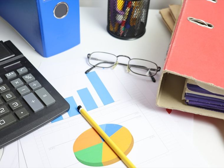Calculator, reading glasses and printed financial charts beside binders of records